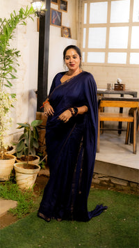 Woman in a dark blue saree standing outdoors with plants and a table in the background