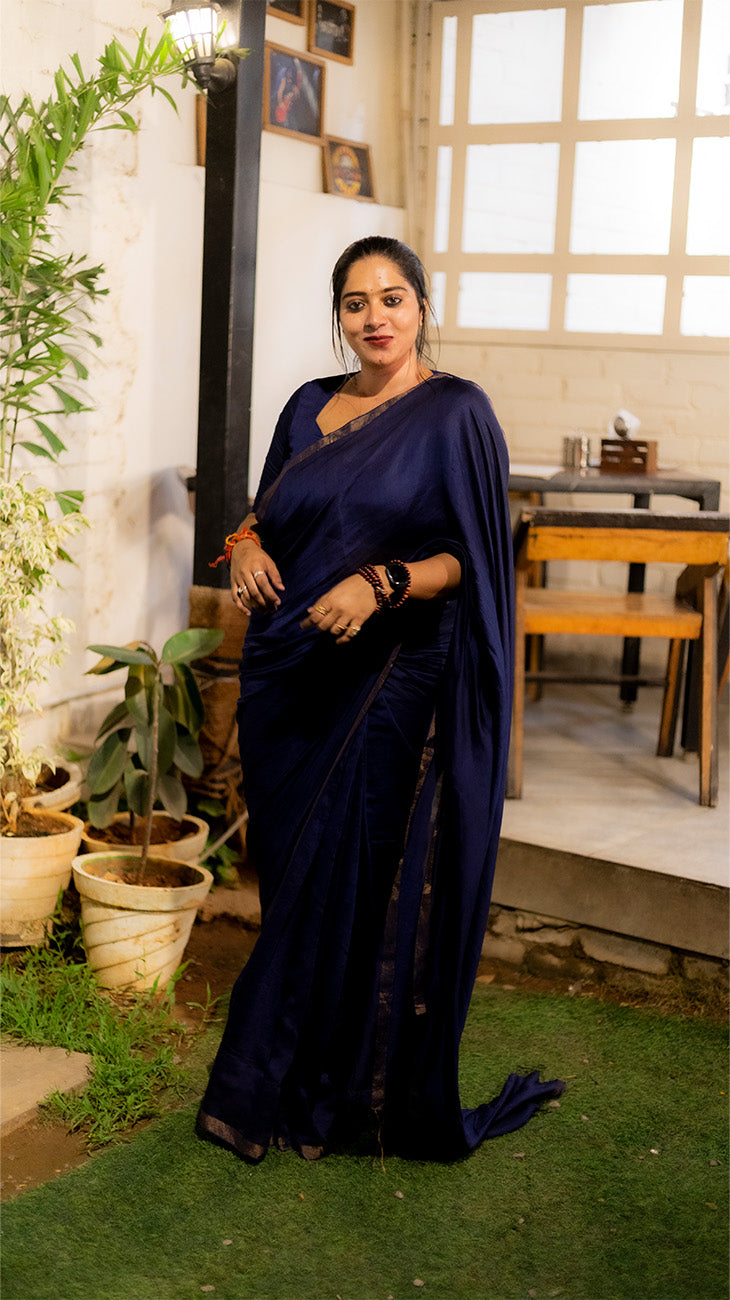 Woman in a dark blue saree standing outdoors with plants and a table in the background