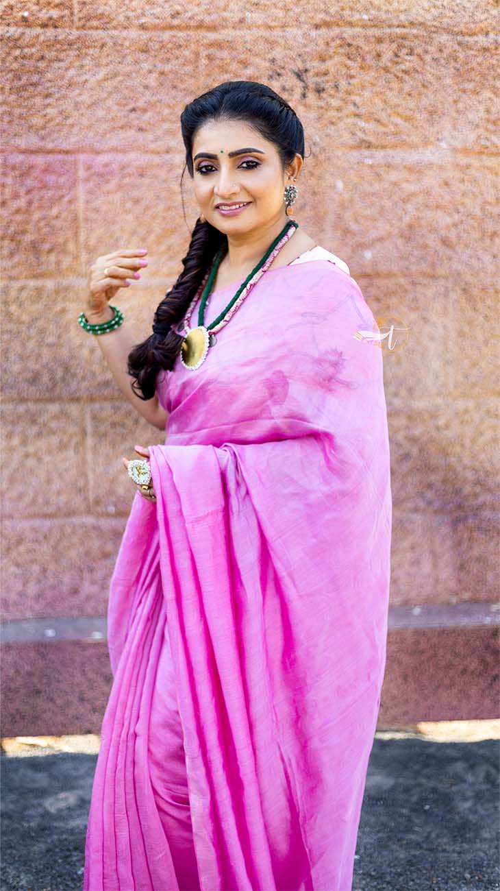 Woman in a pink saree standing against a textured wall.